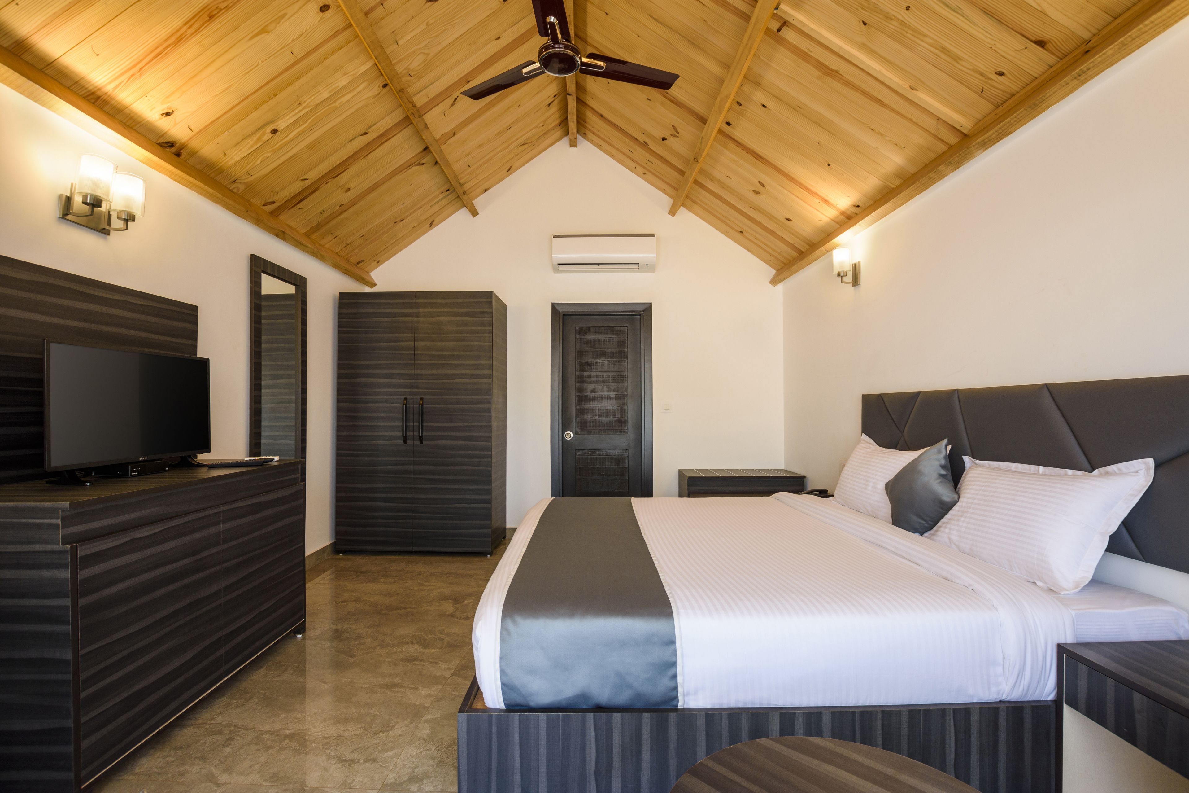 A modern bedroom with a wooden ceiling, a flat-screen TV, a large bed with grey bedding, and a dark-colored wardrobe.