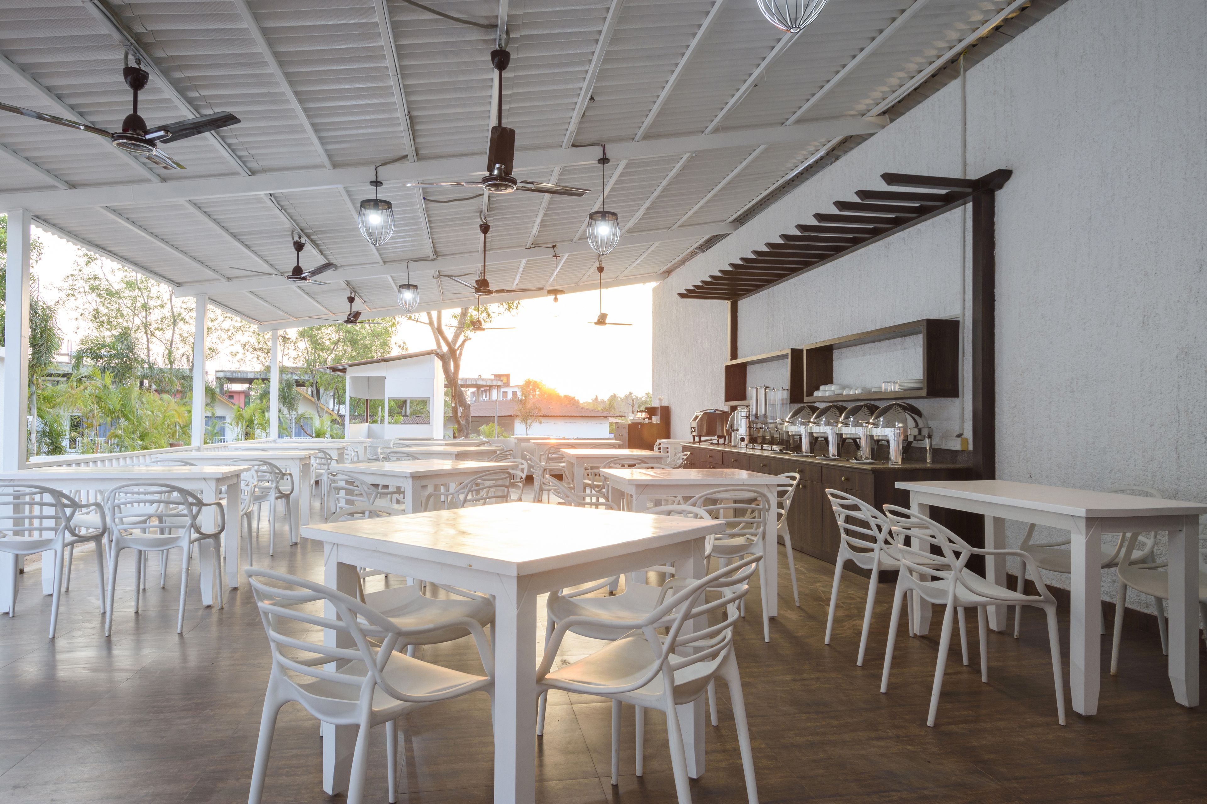 Un vaste espace extérieur aménagé pour les repas, avec des tables et des chaises blanches, des ventilateurs de plafond et un coin bar doté d’un comptoir et d’étagères.