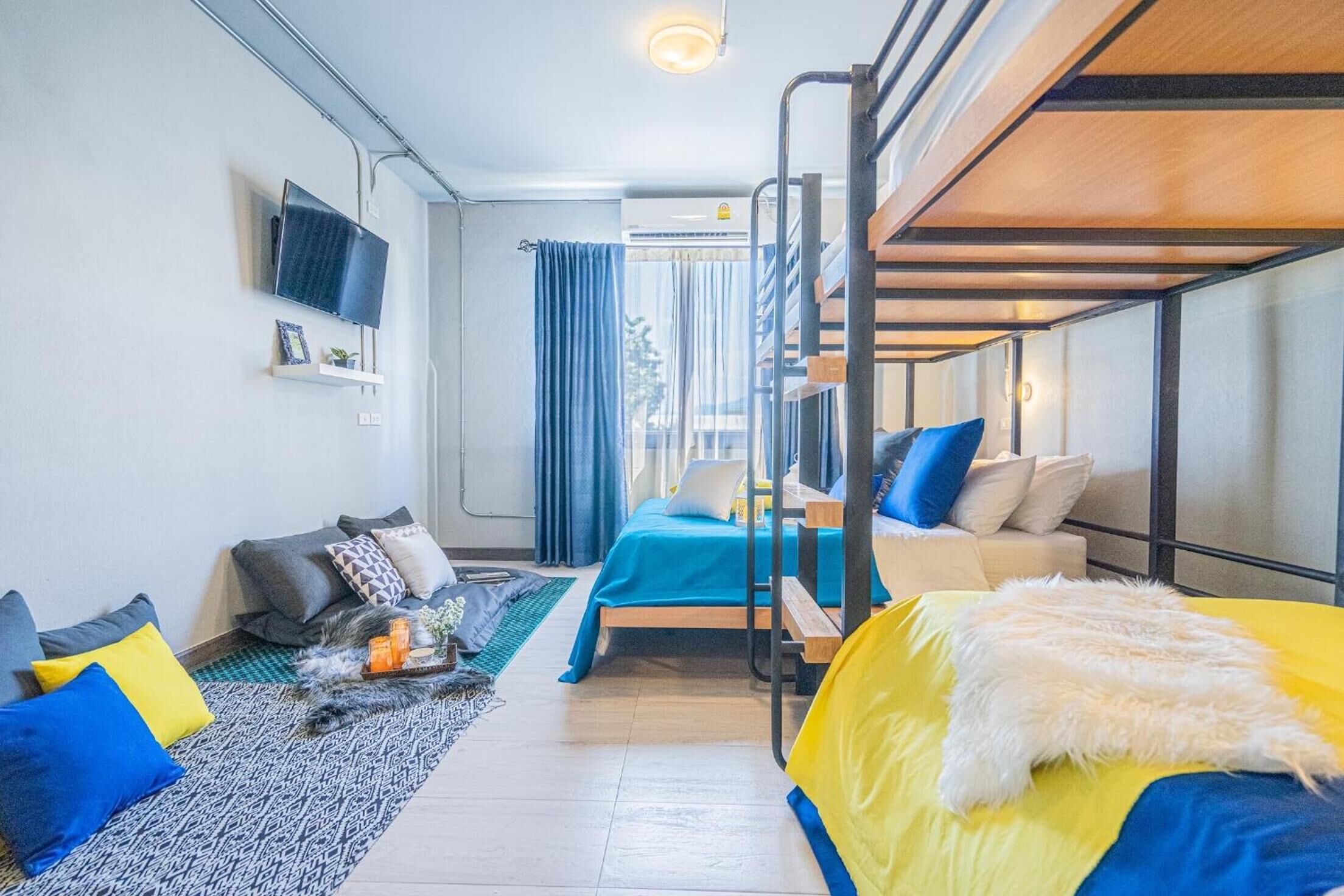 A compact hotel room with bunk beds, a flat-screen TV, a small seating area with cushions, and a window with curtains.
