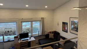 A modern room with a desk, computer, and a black sofa. There are large windows with a view of trees and a radiator under the desk. - Ferienhaus am Stausee Bitburg (Biersdorf am See)