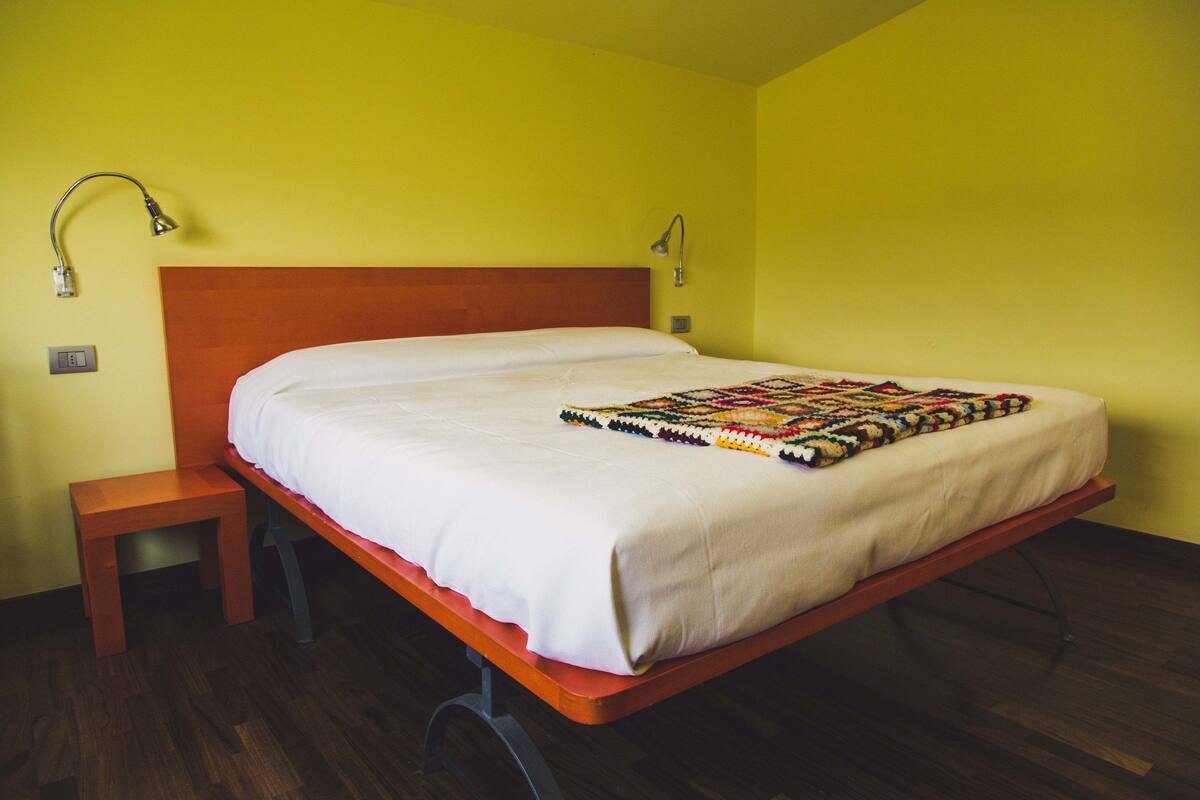 Un lit simple avec une tête de lit en bois et un couvre-lit blanc, accompagné d’une petite table de chevet en bois et d’une courtepointe aux couleurs vives.