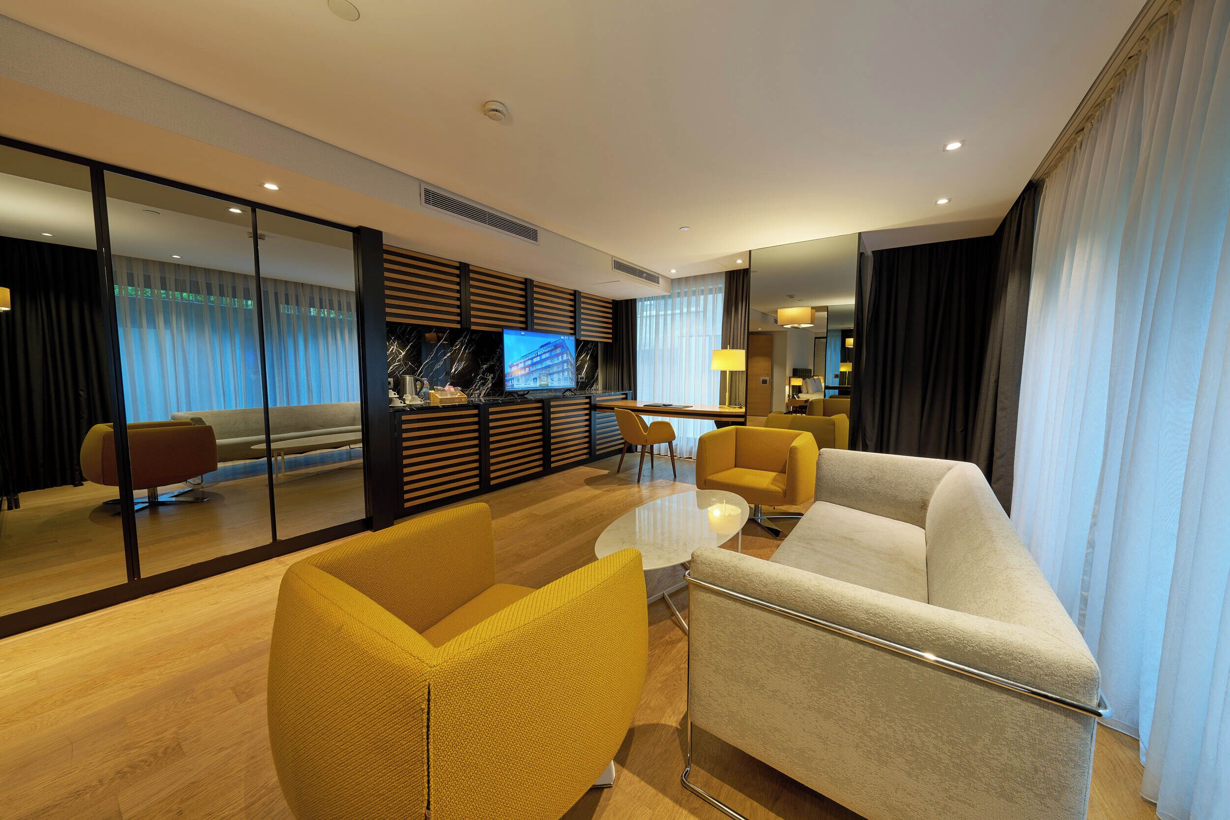 A modern hotel room with a sofa, armchairs, a TV, and a glass partition.