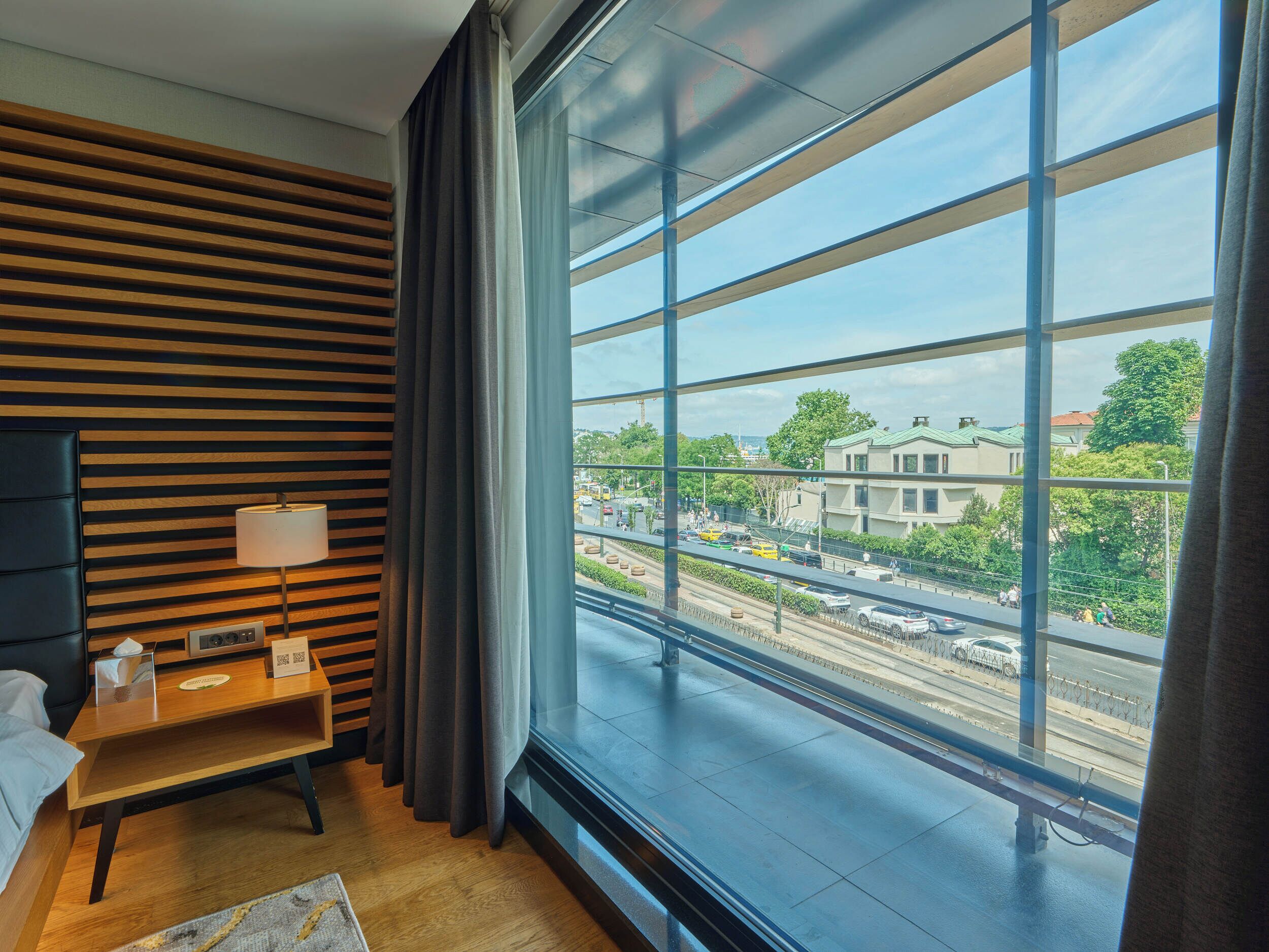 Een moderne hotelkamer met een groot raam, een wand van houten latten, een nachtkastje met een lamp en uitzicht op de stadsplattegrond.