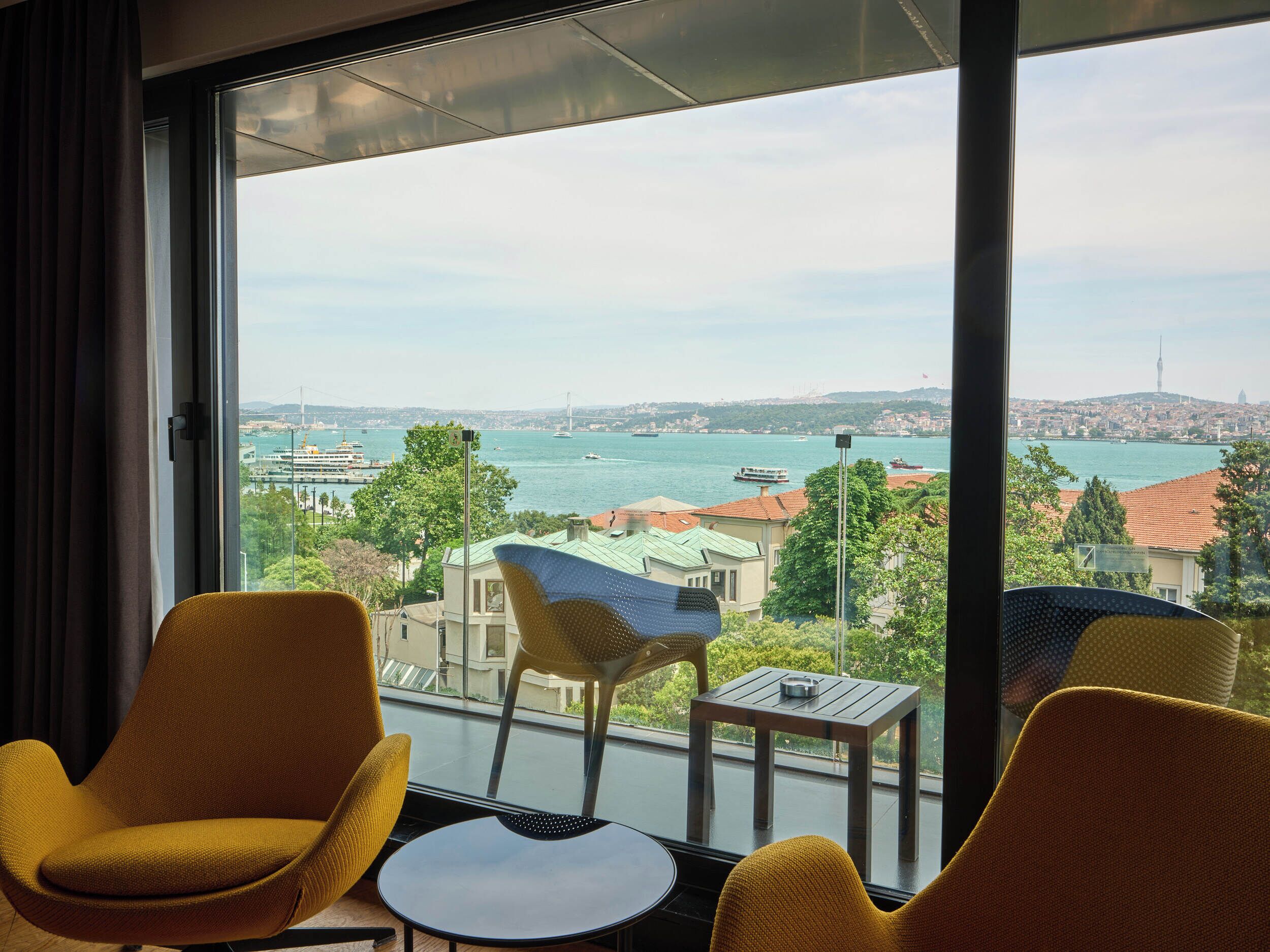 A room with a large window, two yellow chairs, a small table, and a view of a cityscape with a bridge and water.