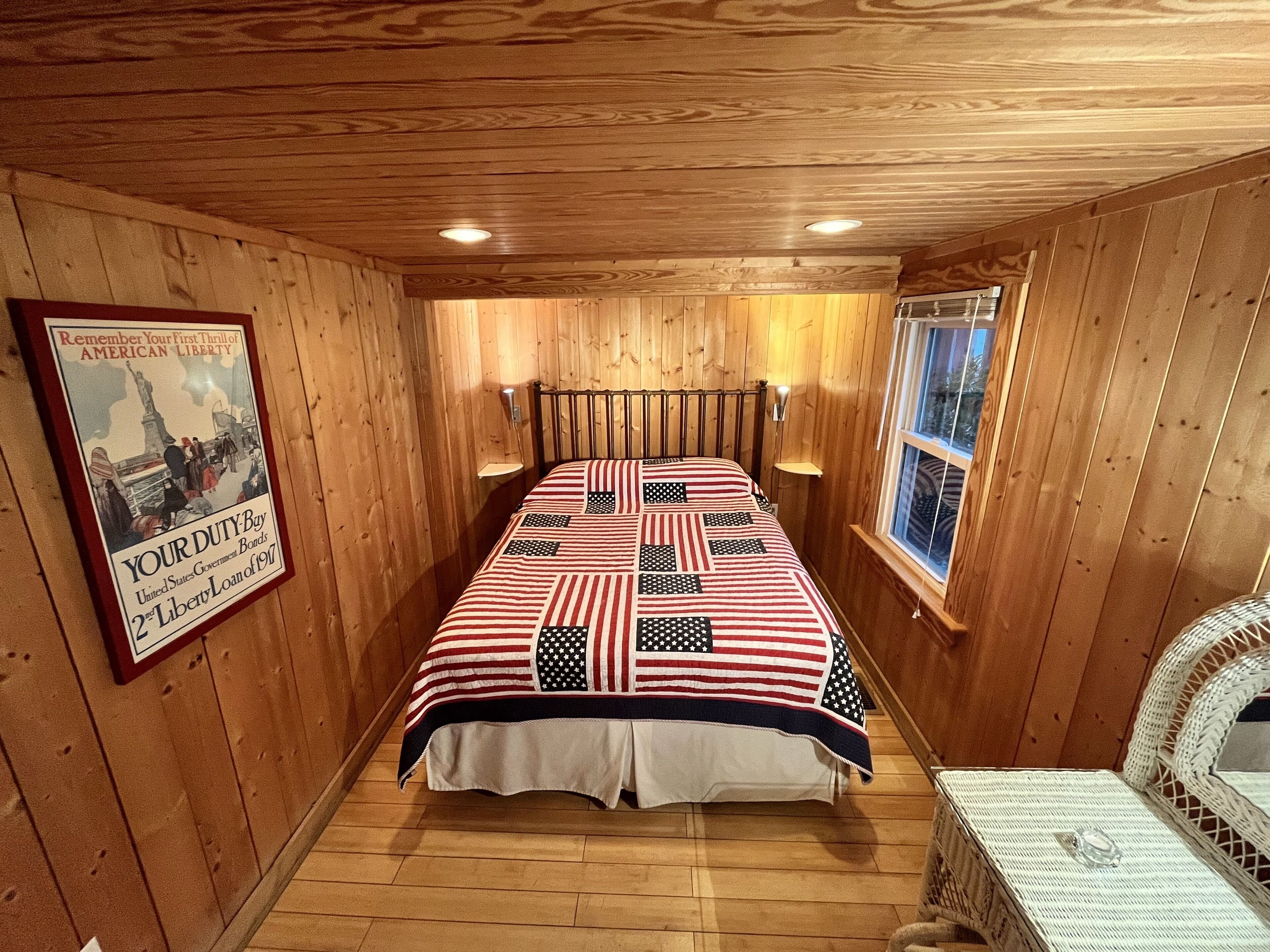 Una habitación con paneles de madera, una cama con diseño de la bandera estadounidense, un cartel histórico enmarcado y una ventana con vistas.