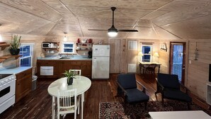 Una cocina compacta con armarios de madera, un microondas y un refrigerador. Cuenta con una zona de comedor con una mesa y sillas, y un ventilador de techo.