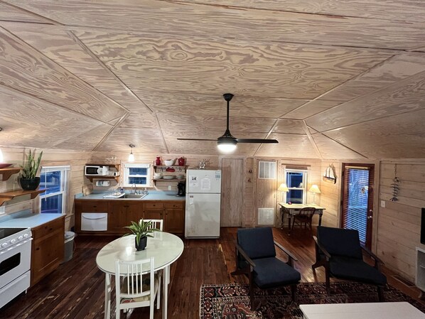 A compact kitchen with wooden cabinets, a microwave, and a refrigerator. There is a dining area with a table and chairs, and a ceiling fan.