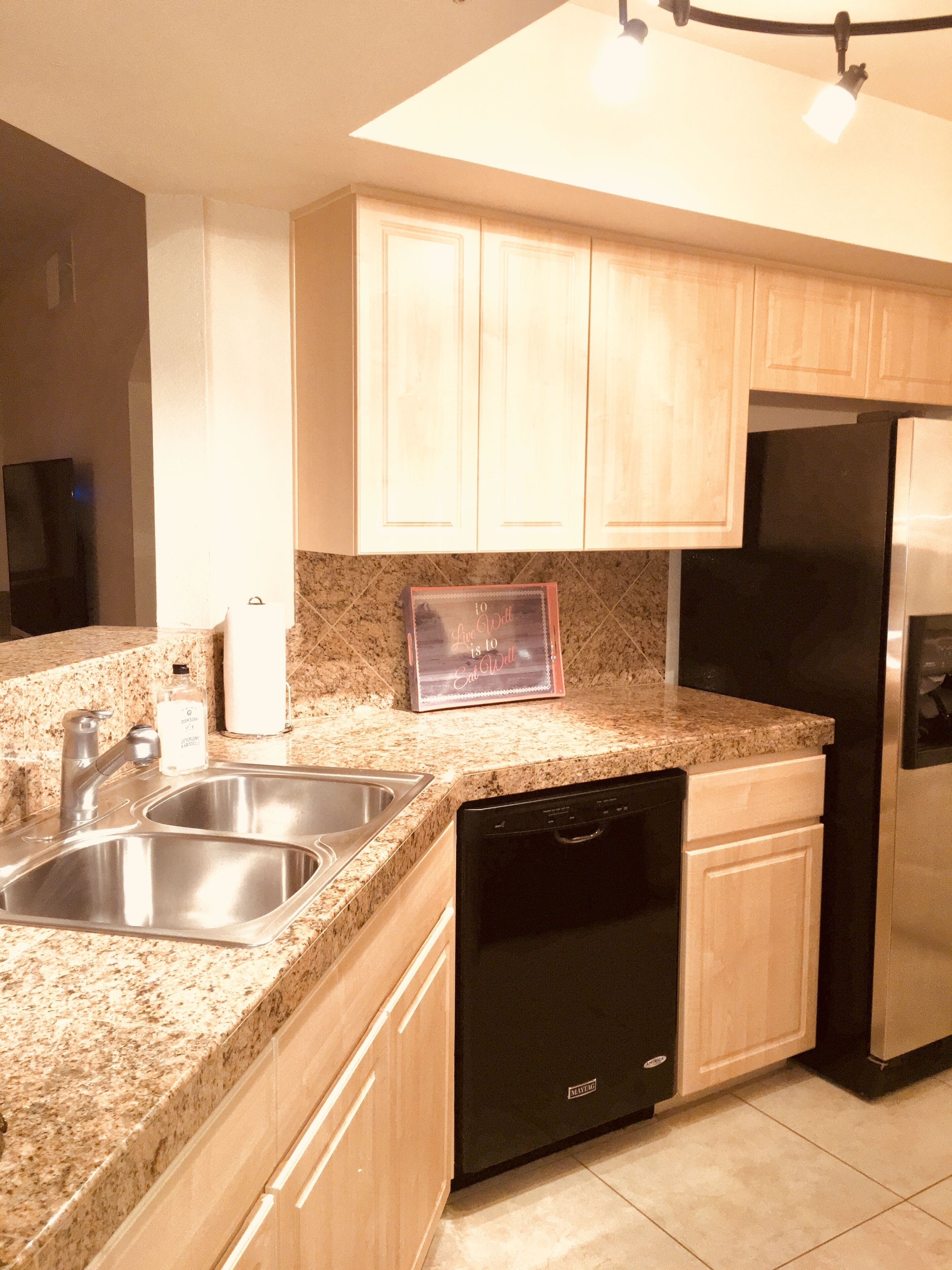 Una cocina con encimeras de granito, pileta de acero inoxidable, lavavajillas negro y muebles de madera.