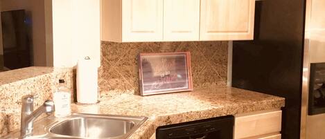 Una cocina con encimeras de granito, pileta de acero inoxidable, lavavajillas negro y muebles de madera.