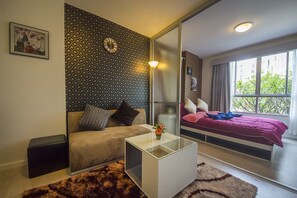 A modern room with a sofa, a glass coffee table, a bed with a red bedspread, a patterned wall, and a window with a view of greenery.