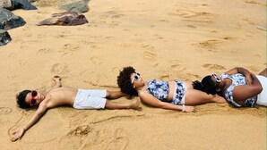 A beach with three people lying on the sand, a rocky shoreline, and a clear sky. - Veivaag Lodge (Sekondi-Takoradi)