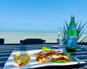 A meal with a salad and omelette on a white plate, a bottle of Pellegrino water, and a glass on a table with a view of the ocean. - Veivaag Lodge (Sekondi-Takoradi)