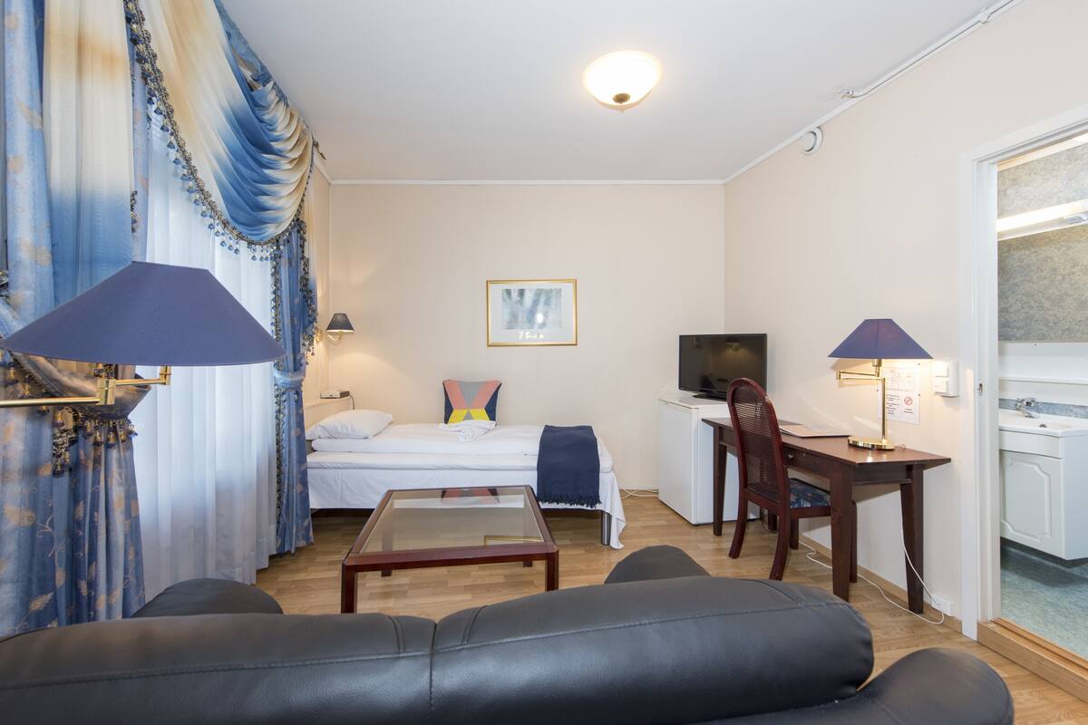 A compact hotel room with a bed, a desk, a television, and a seating area with a couch and a table.