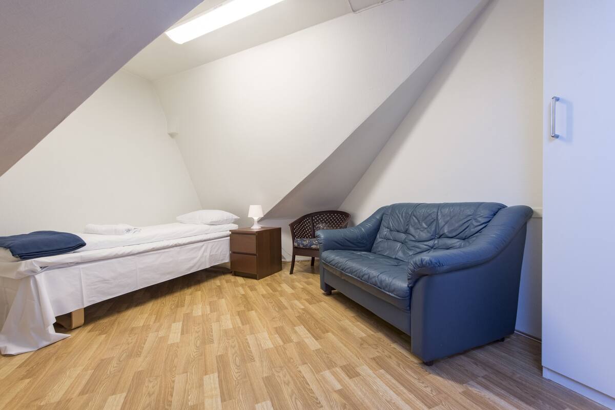 A small, triangular room with a bed, a blue chair, a nightstand, and a lamp.