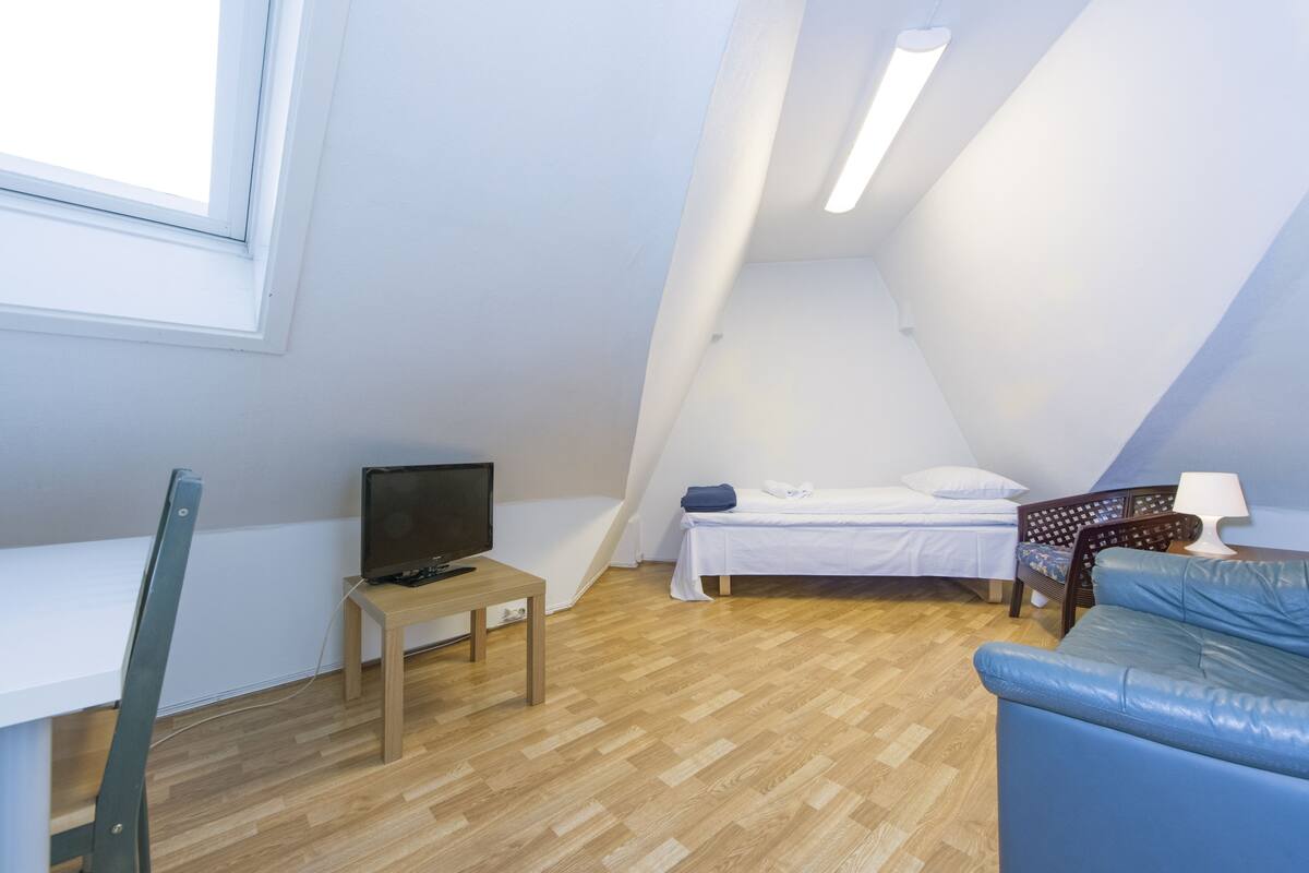 A small, well-lit room with a bed, a desk with a computer, a blue sofa, and a window.