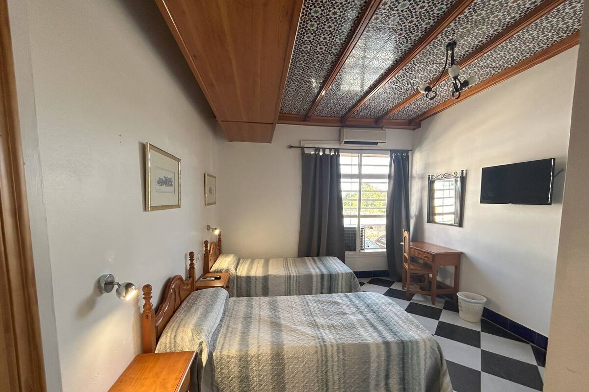 A bedroom with two beds, a desk, a chair, a TV, and a window with curtains.