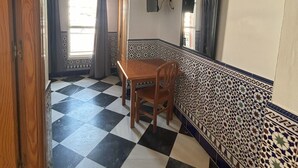 A room with a checkered floor, a wooden table, and chairs. There is a window with curtains, a wall-mounted mirror, and a patterned ceiling.