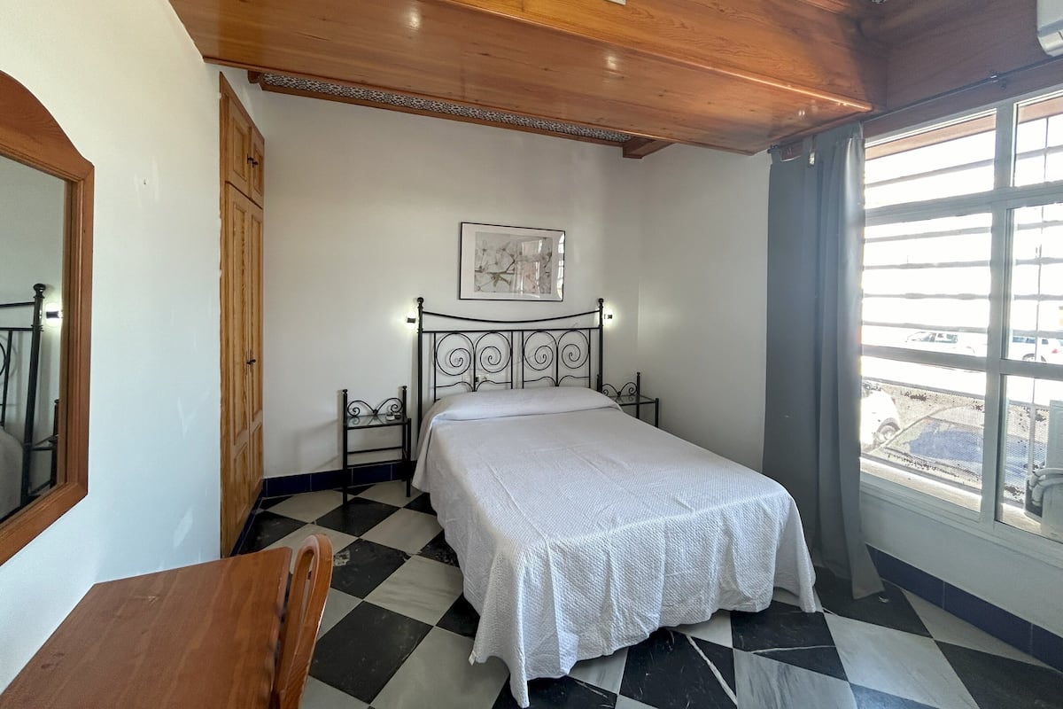 A bedroom with a bed, a wooden chair, a window with a view, and a ceiling vent.