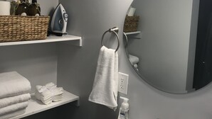 Une salle de bain avec un lavabo blanc, un miroir rond, un porte-serviettes et des étagères contenant des articles de toilette et un fer à repasser.