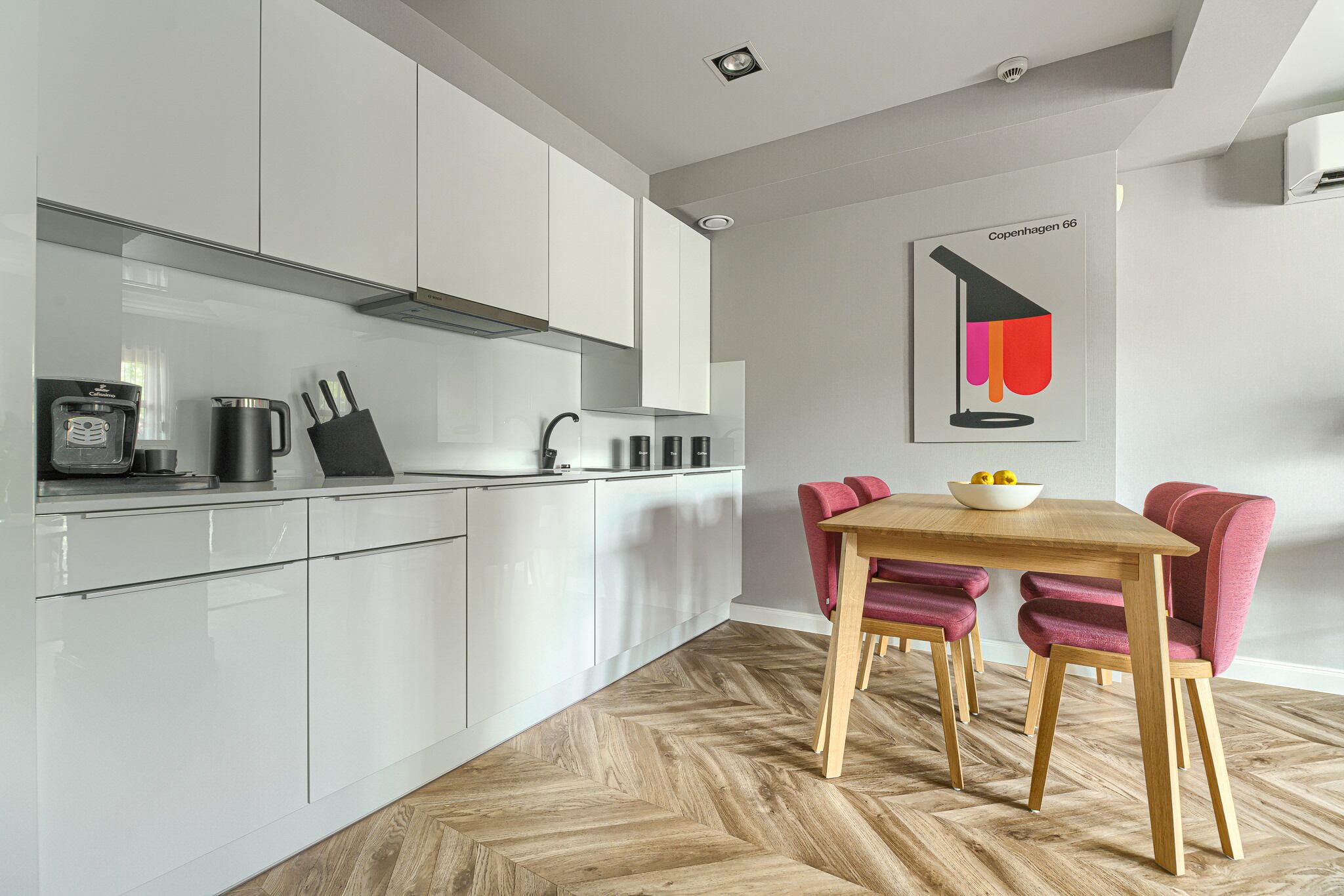 A modern kitchen with white cabinets, a wooden herringbone floor, and a dining area with a wooden table and red chairs.