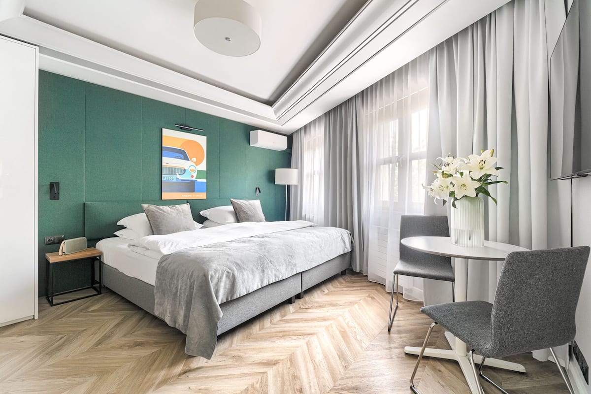 A modern hotel room with a large bed, a small table with a chair, a vase of flowers, and a painting on the wall.