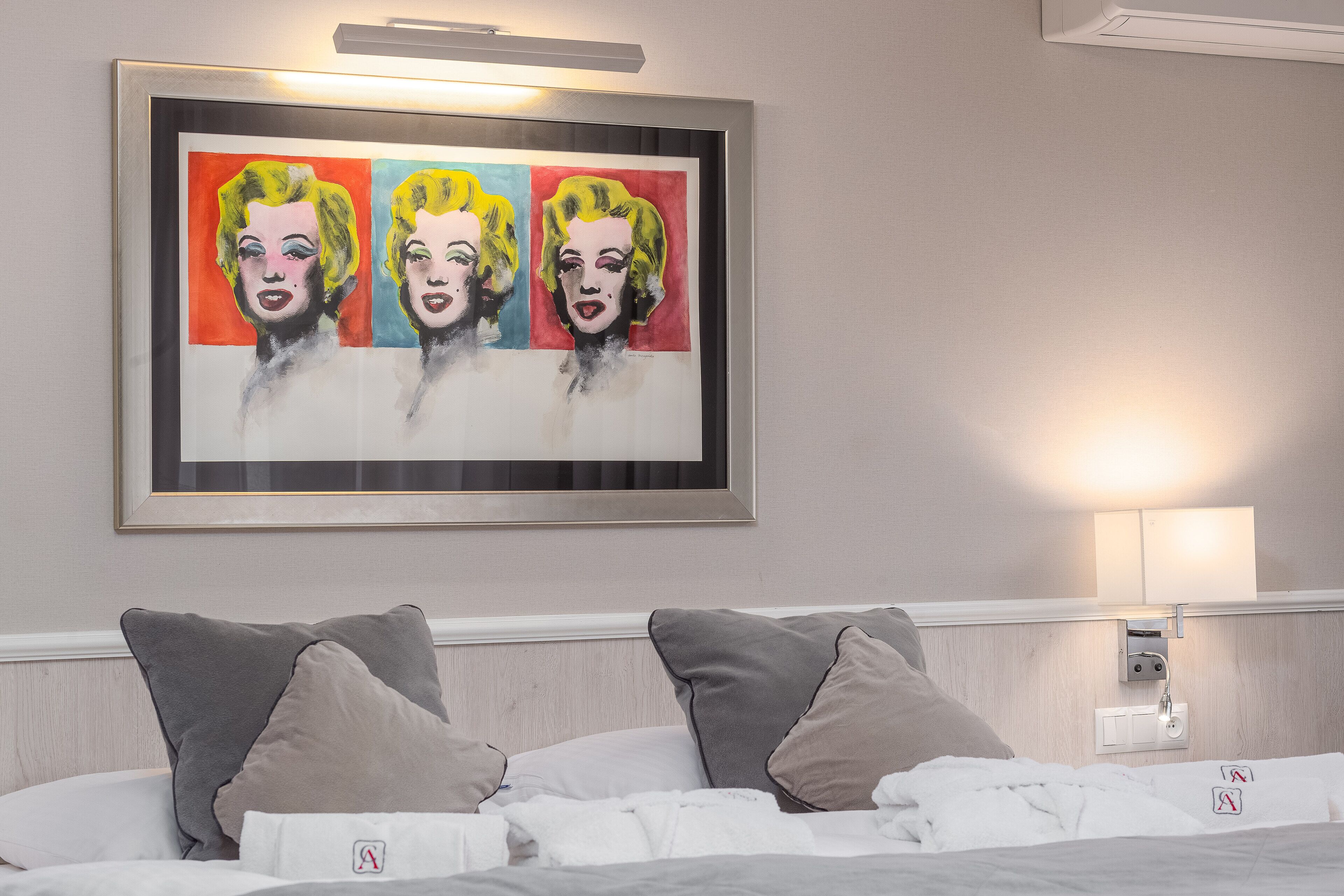 A hotel room with a bed, pillows, a framed artwork above the bed, and a wall-mounted light.