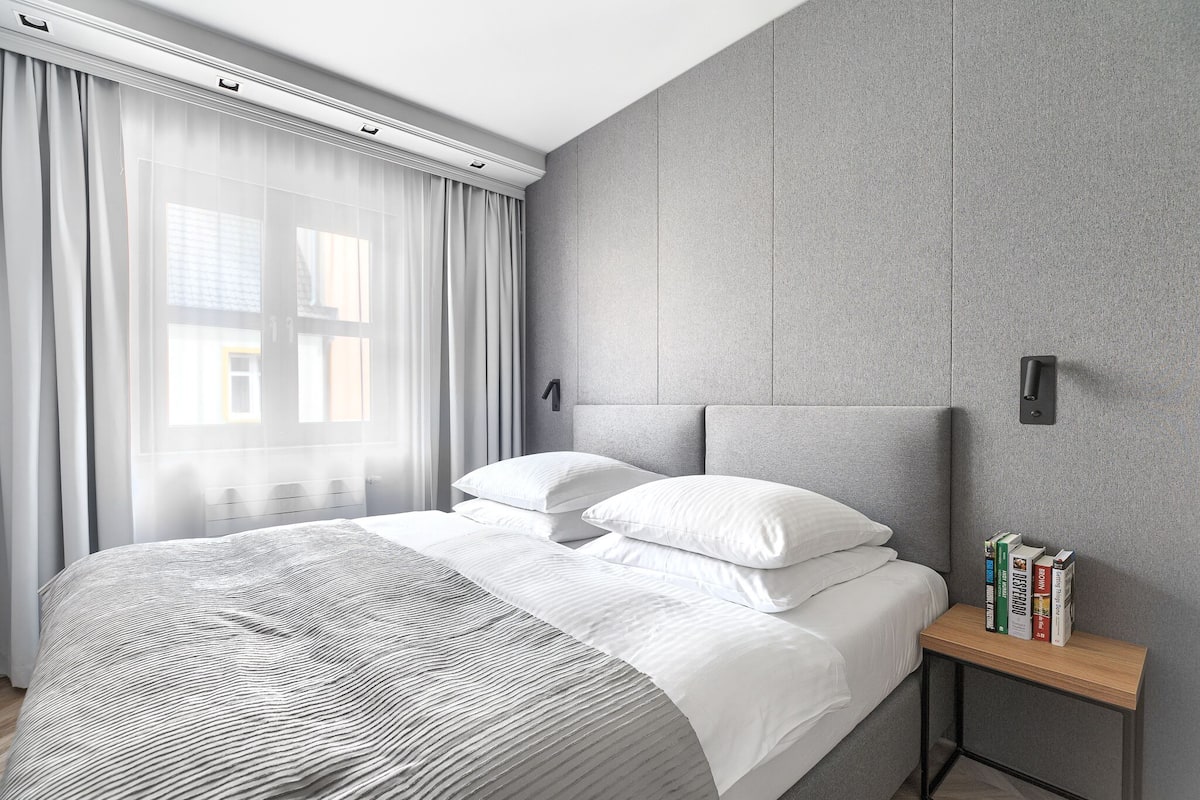 A modern bedroom with a large bed, a bedside table with books, and a view of the outside through sheer curtains.
