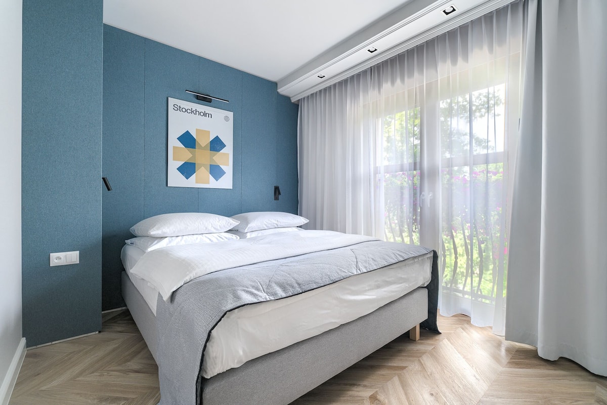 A bedroom with a bed, a wall-mounted air conditioning unit, and a window with sheer curtains.