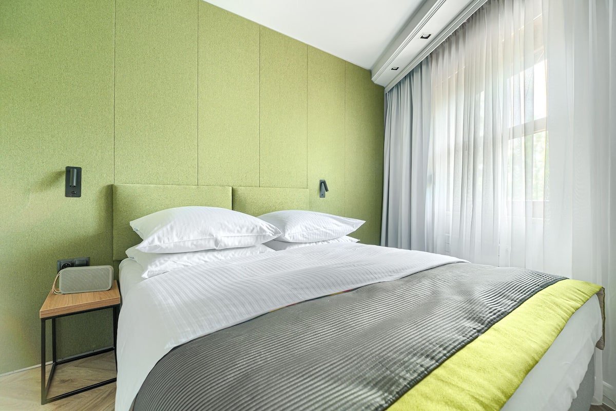 A neatly made bed with white linens and a grey and yellow striped blanket, a wooden bedside table with a speaker, and a green wall with a window covered by sheer curtains.