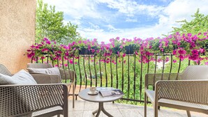 Un espace extérieur aménagé avec des meubles en osier et une table, donnant sur un jardin où fleurissent des fleurs roses et qui est délimité par une clôture en métal.