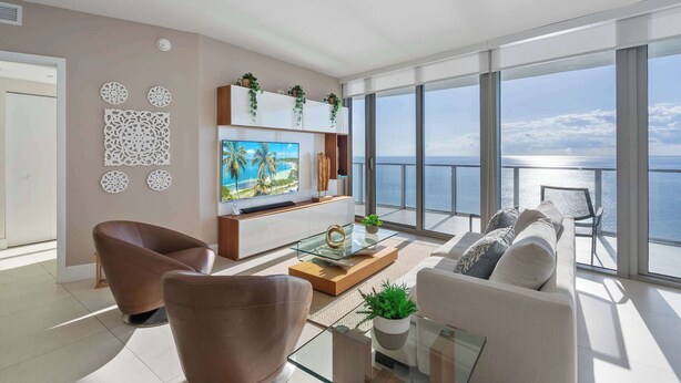 En moderne stue med stort tv, behagelige siddepladser, et glasbord og en balkon med havudsigt.
