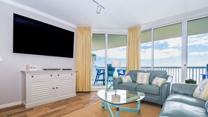 A living room with a flat-screen TV, a glass coffee table, a sofa, and a sliding glass door leading to a balcony. - Hardwood flooring throughout updated, tropical condo! 🌺 (Destin)