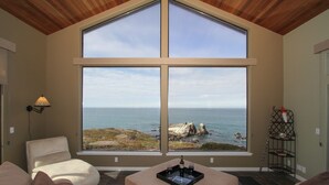 A room with a large window overlooking the ocean, a cushioned seating area, and a small table with a bottle and glasses. - Private, Oceanfront Views, Starlink WiFi, Timber Cove (Jenner)