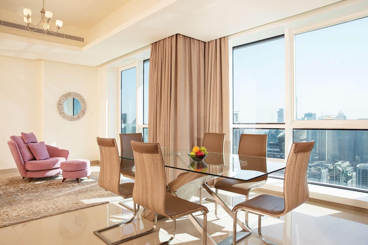 A modern living room with a glass-top dining table, brown leather chairs, a pink armchair, and a city view through large windows.