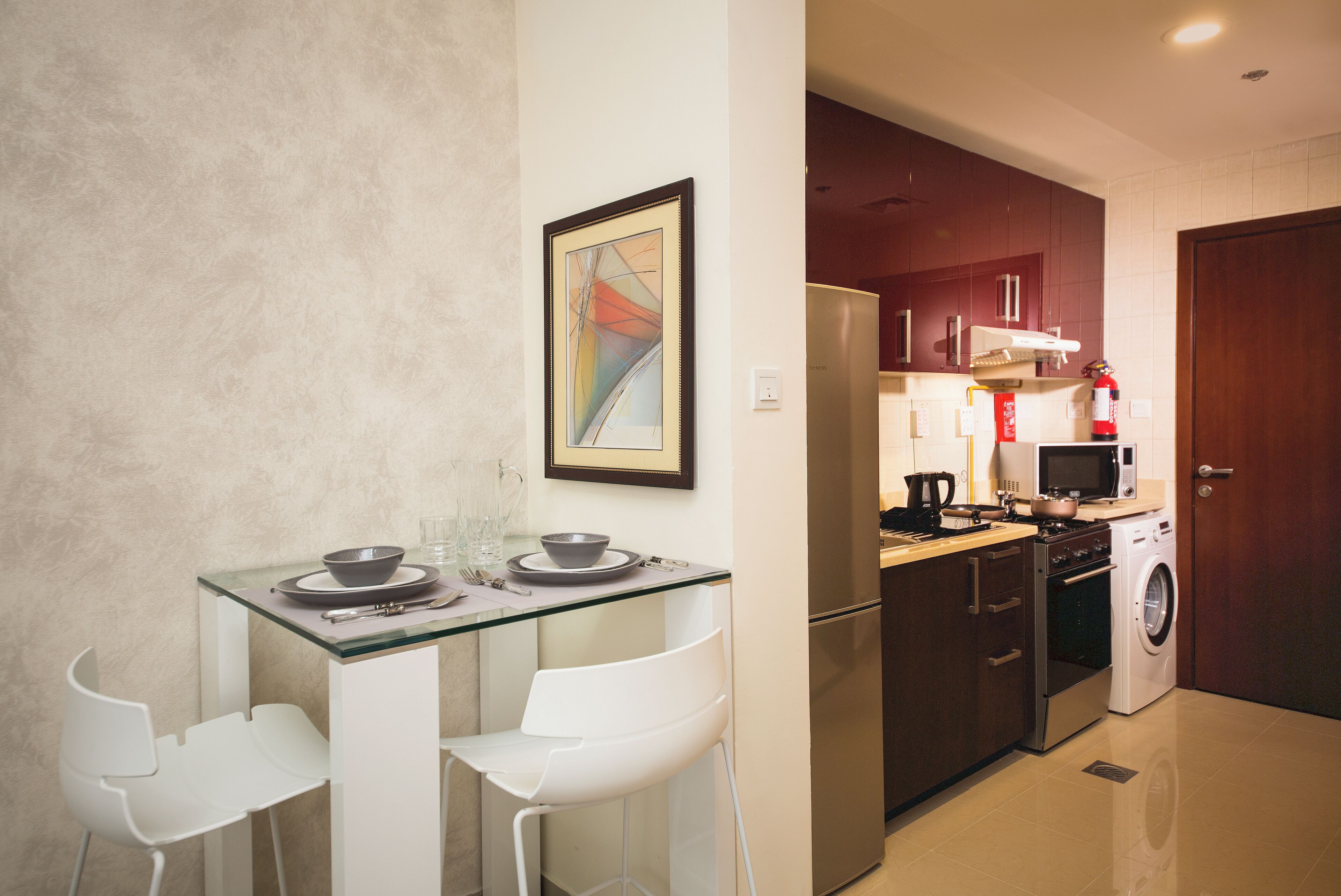 A compact kitchen with a refrigerator, microwave, stove, and washing machine. There is a glass table with two white chairs and a framed picture on the wall.