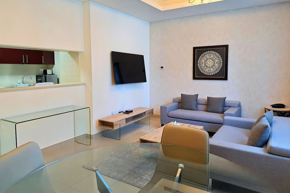 A modern living room with a sofa, a coffee table, a glass dining table, a flat-screen TV mounted on the wall, and a kitchen area with cabinets and a microwave.