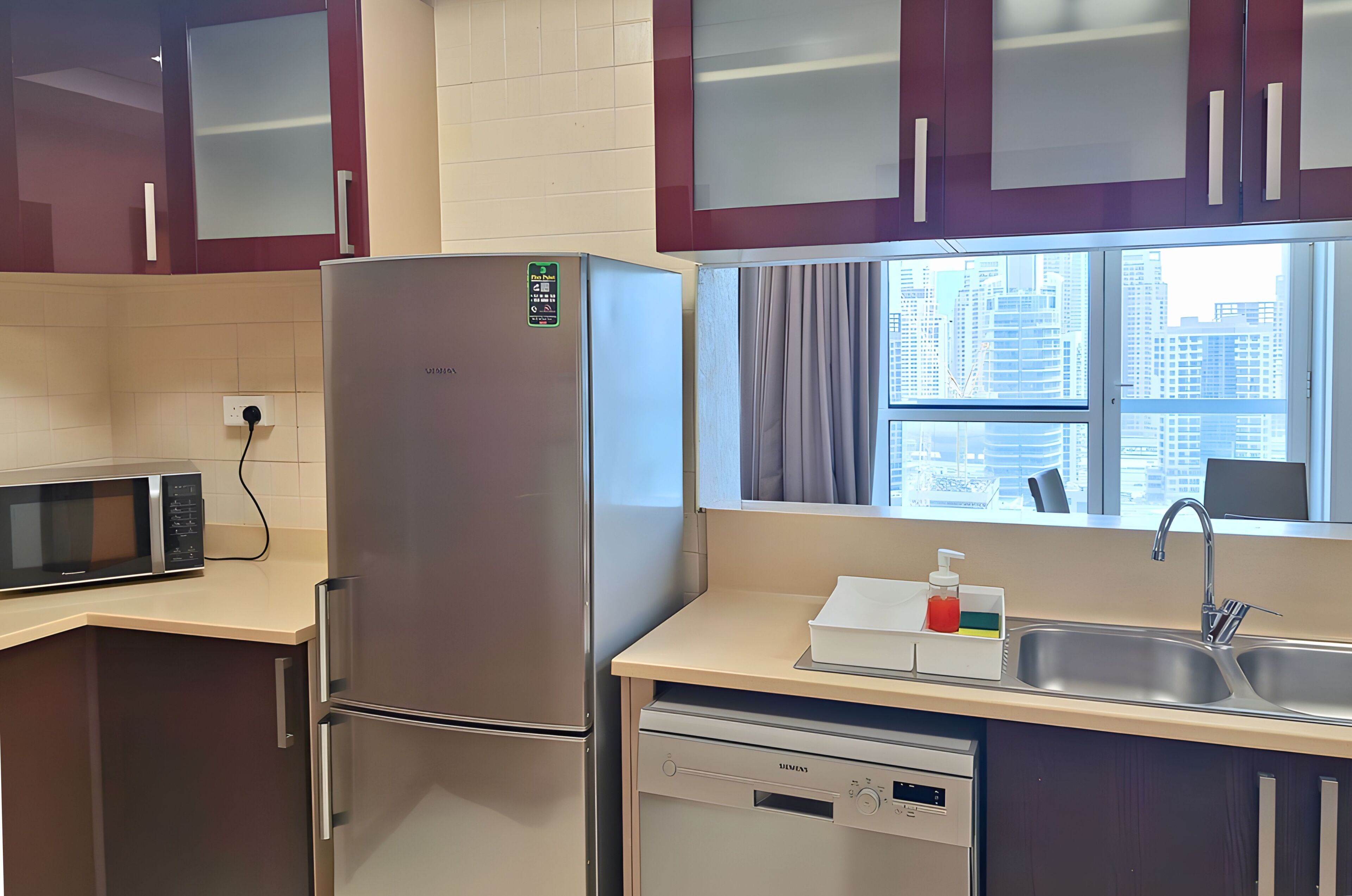 A modern kitchen with a stainless steel refrigerator, a microwave, a dishwasher, and a window with a city view.