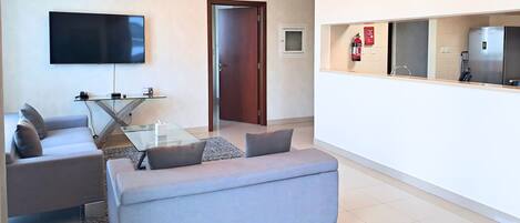 A modern living room with a grey sofa, a glass coffee table, a flat-screen TV, and a kitchen area with a fridge and microwave.