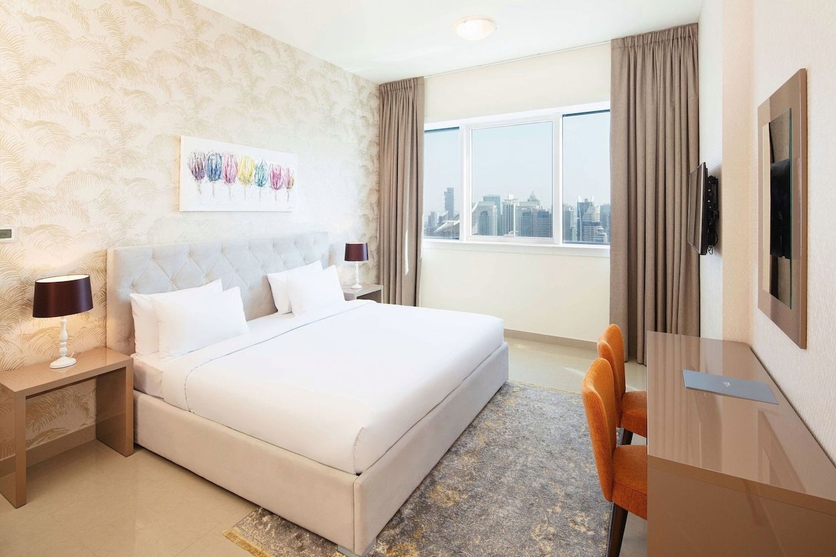 A hotel room with a large bed, a desk, a television, and a city view.