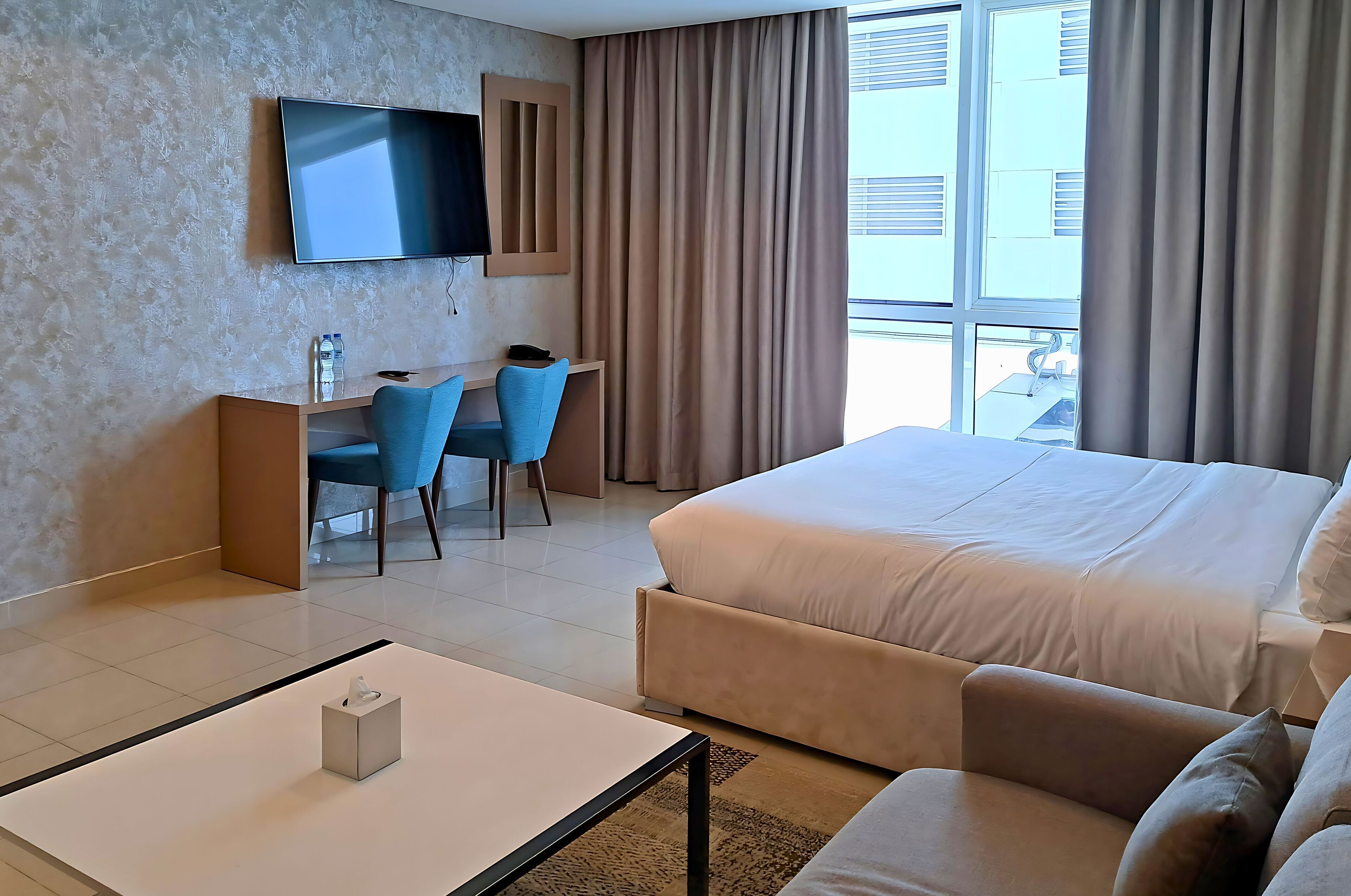 Een hotelkamer met een bed, een bureau met stoel, een aan de muur bevestigde televisie en een raam met gordijnen.