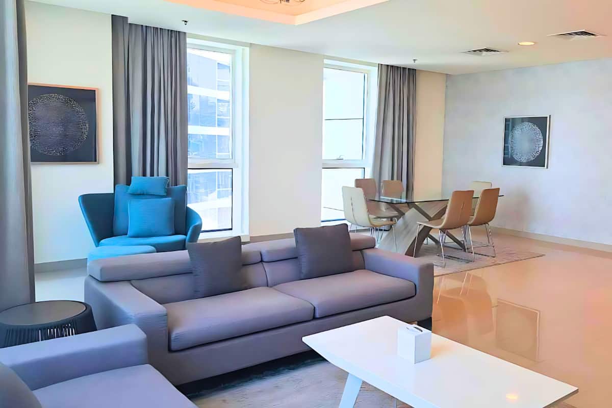A modern living room with a grey sofa, a blue armchair, a dining table with chairs, and large windows with curtains.