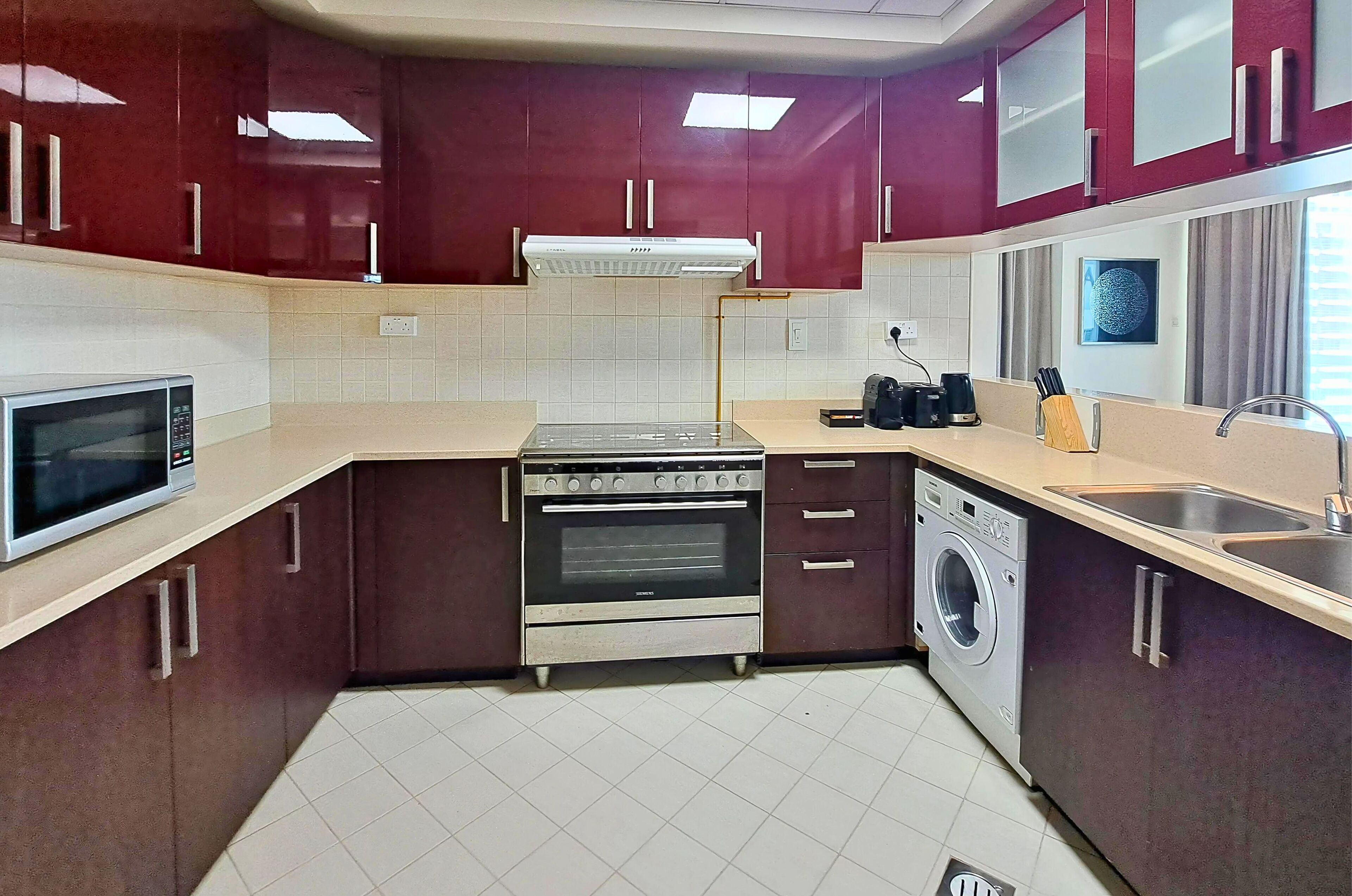 A compact kitchen with a microwave, oven, and washing machine, featuring dark red cabinets and a tiled backsplash.