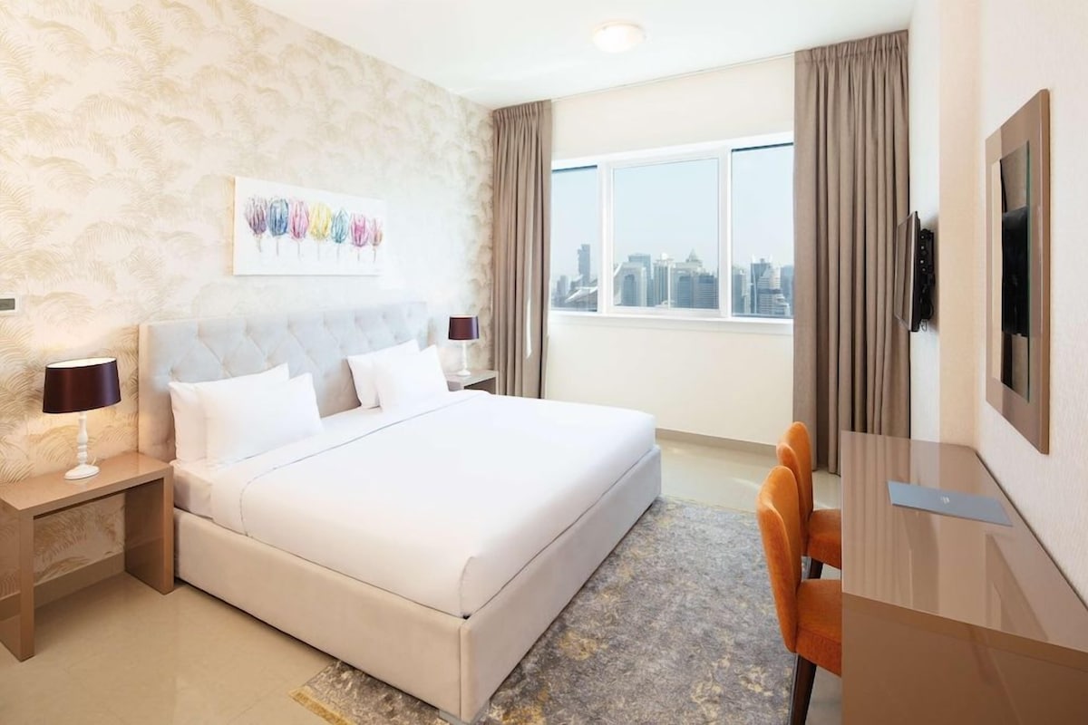 A hotel room with a large bed, a desk with a chair, a television, and a city view.