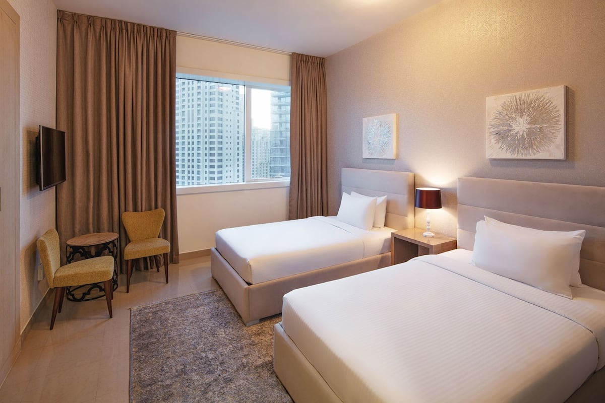 A hotel room with two beds, a TV, a chair, and a window with a city view.