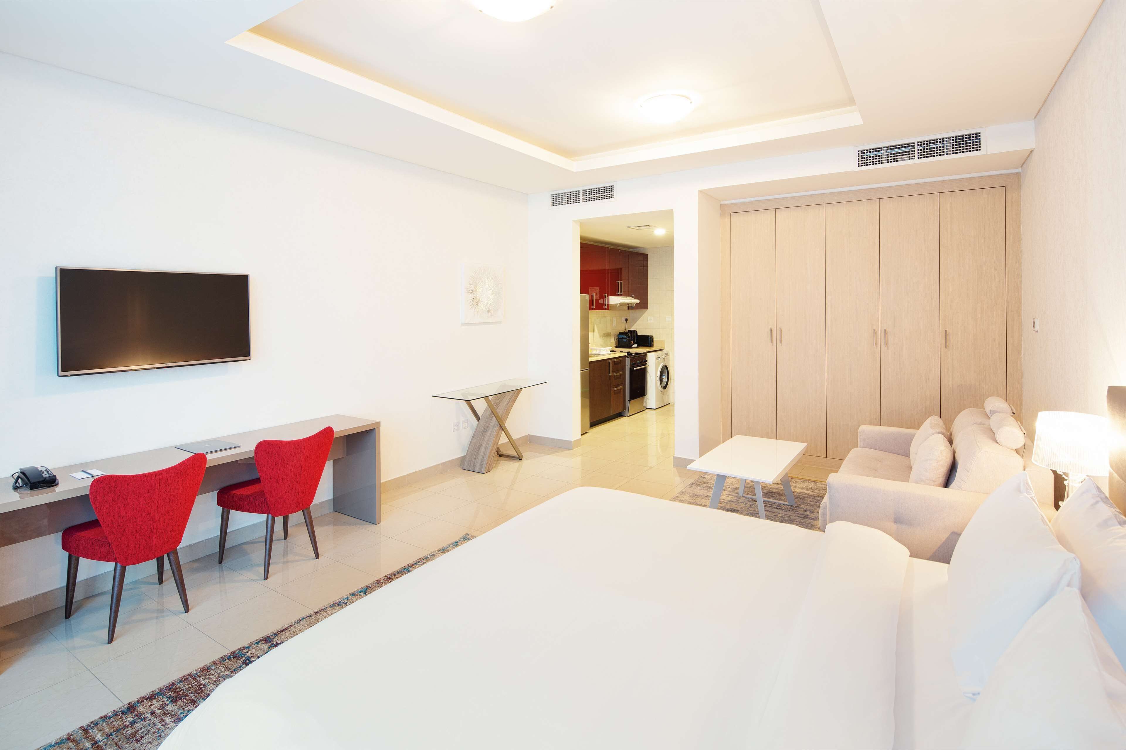 Una habitación de hotel moderna con una cama grande, un escritorio con dos sillas rojas, un televisor y una kitchenette al fondo.