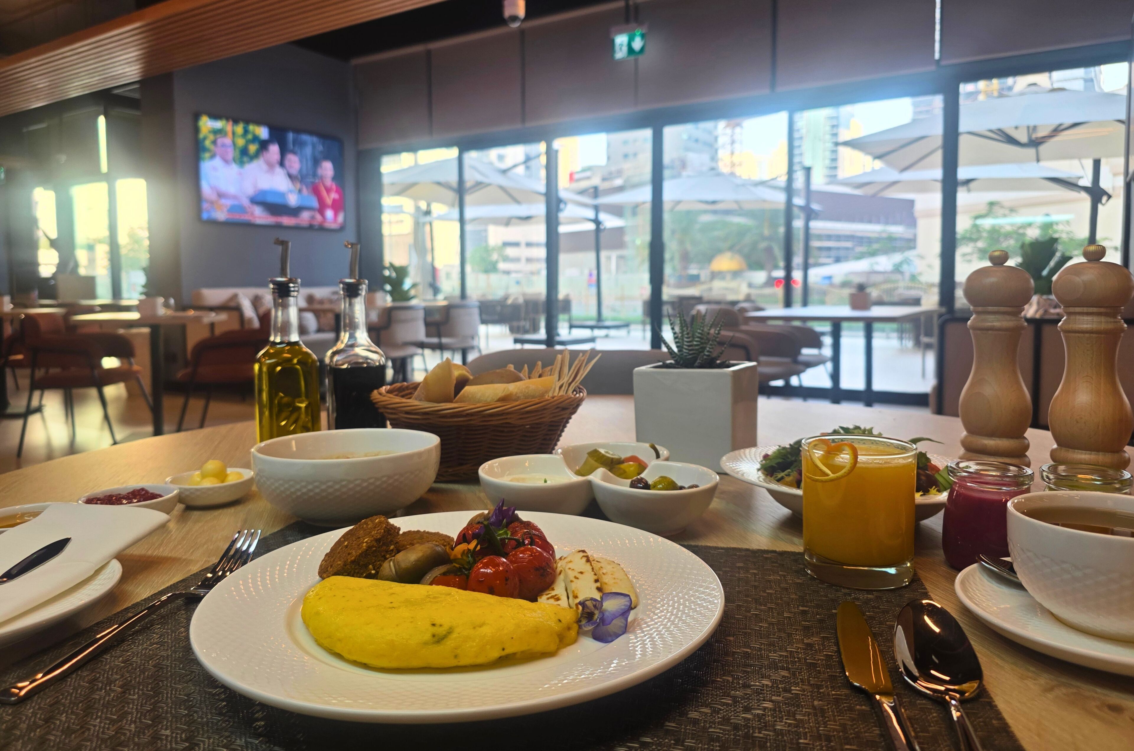 Un ambiente de desayuno con platos de comida, un televisor al fondo y vistas al exterior a través de amplias ventanas.