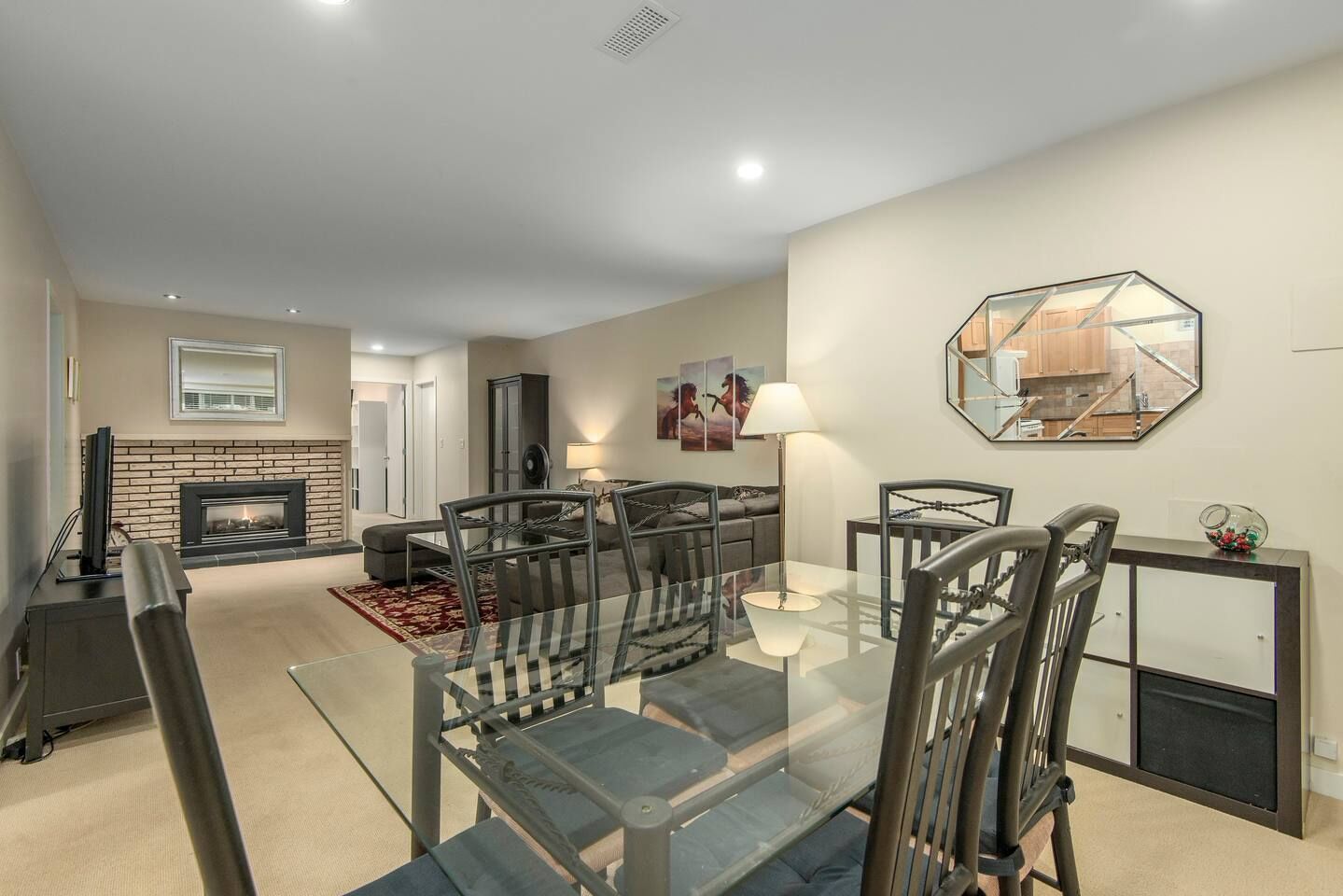A modern living room with a glass dining table, grey upholstered chairs, a fireplace, and a large mirror on the wall.