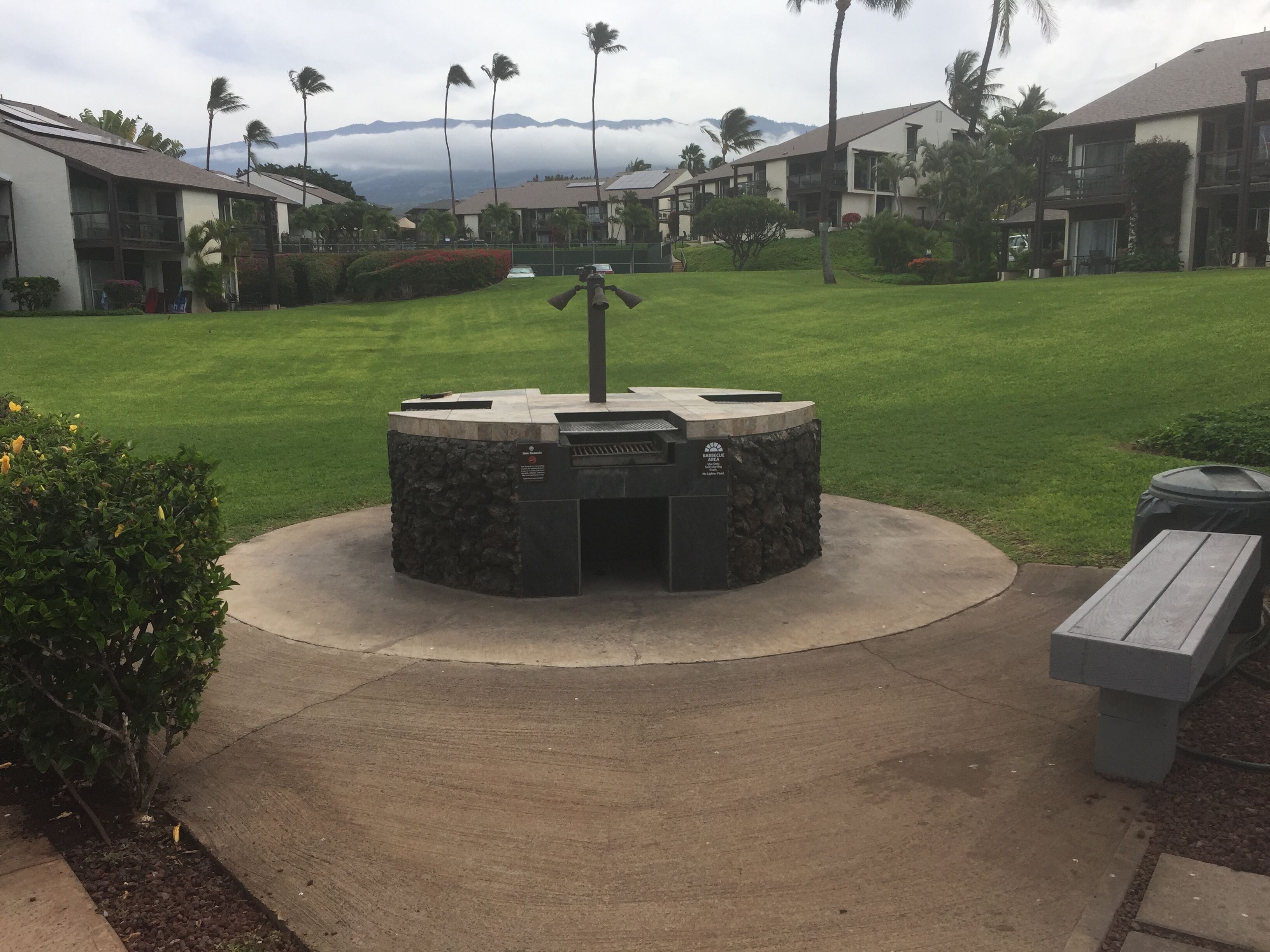 A circular outdoor grill area with a stone base, surrounded by a concrete path, set in a well-maintained grassy area with palm trees and a mountain backdrop.