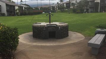 A circular outdoor grill area with a stone base, surrounded by a concrete path, set in a well-maintained grassy area with palm trees and a mountain backdrop.