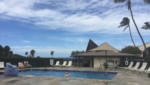 Un área de piscina con sillones de descanso, una zona de estar techada y palmeras.